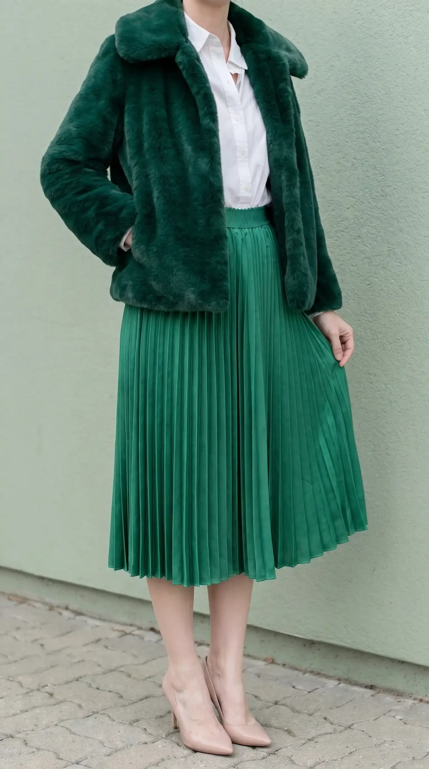 Woman in forest green faux fur jacket with white shirt and green pleated midi skirt and nude pumps