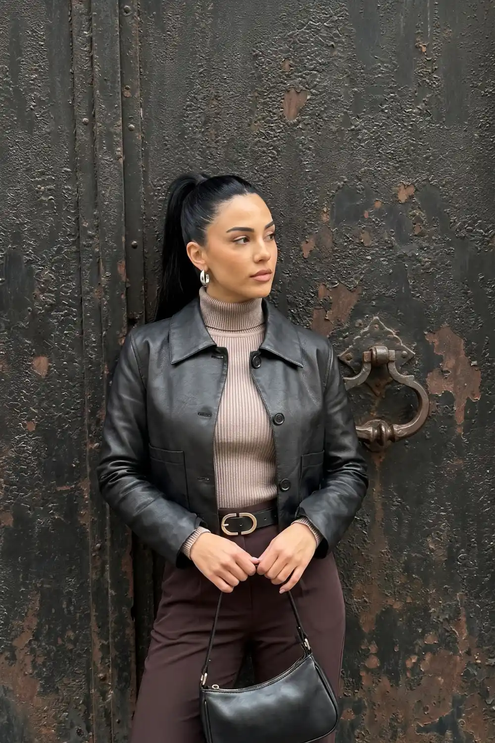 Woman wearing black cropped button front leather jacket with taupe turtleneck and dark brown belted trousers