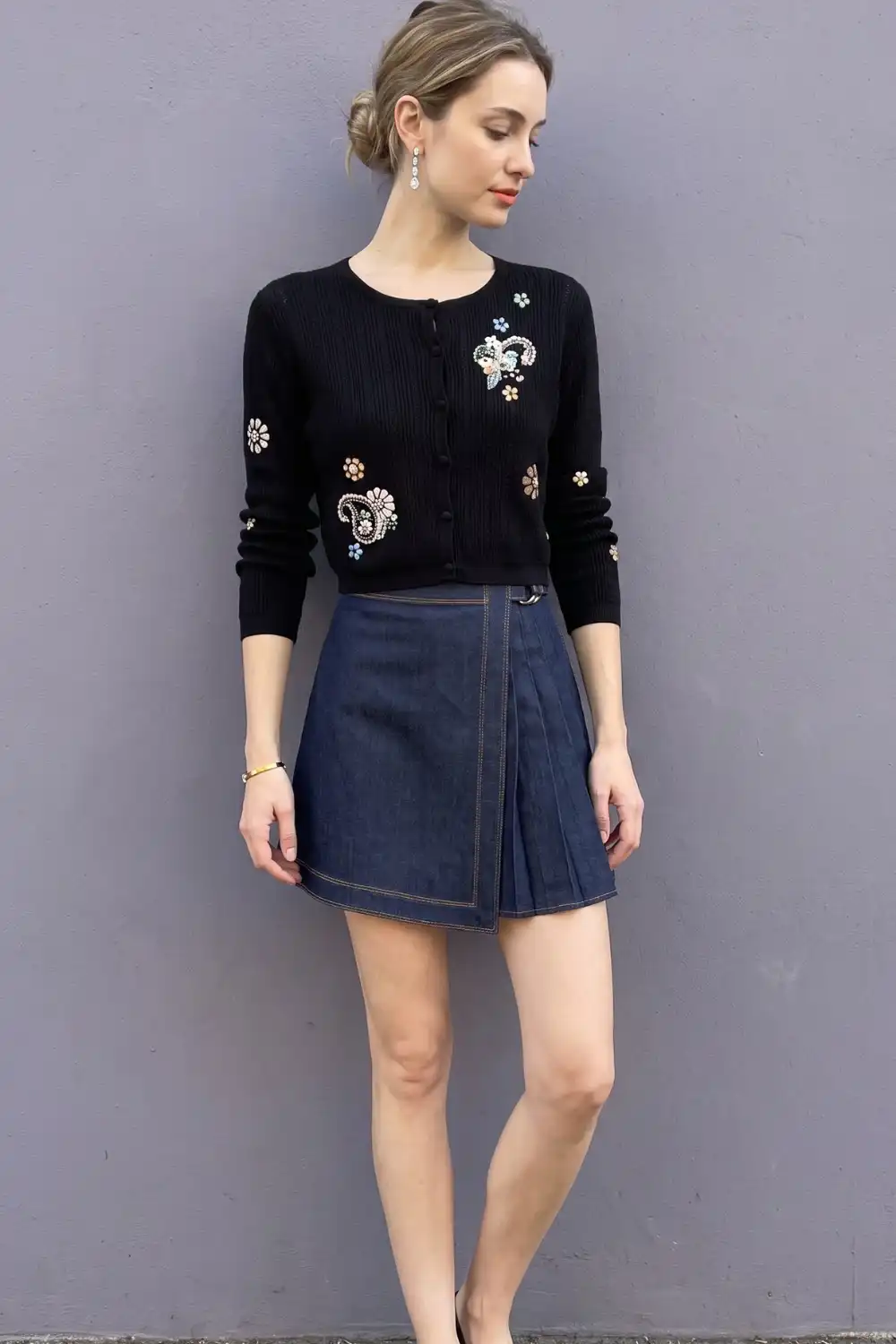 Woman in black embellished floral knit cardigan with dark blue denim pleated wrap mini skirt