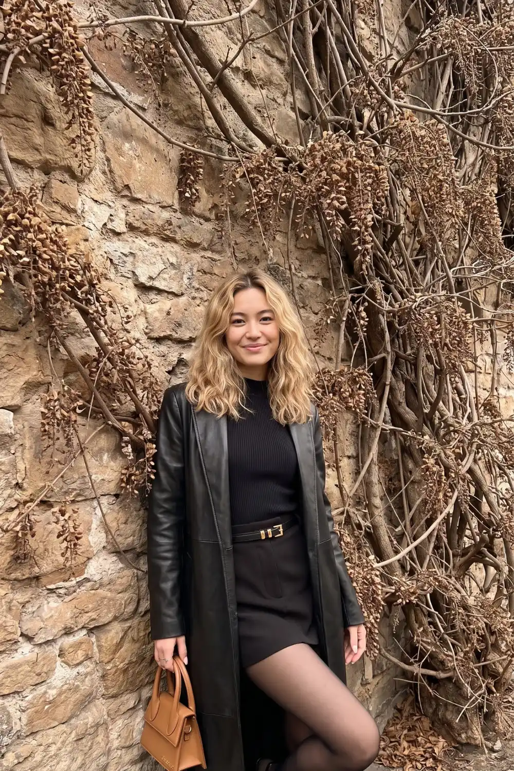 Woman wearing black longline leather coat with ribbed turtleneck belted mini skirt and black tights