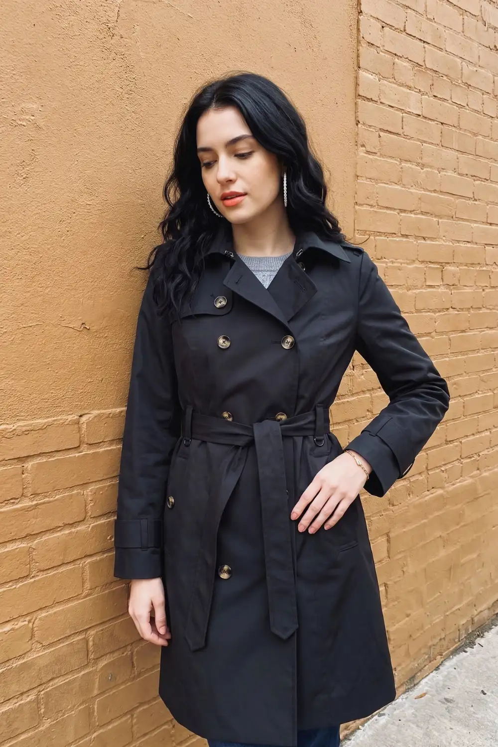 Woman in black double-breasted belted midi trench coat with grey knit sweater and dark jeans