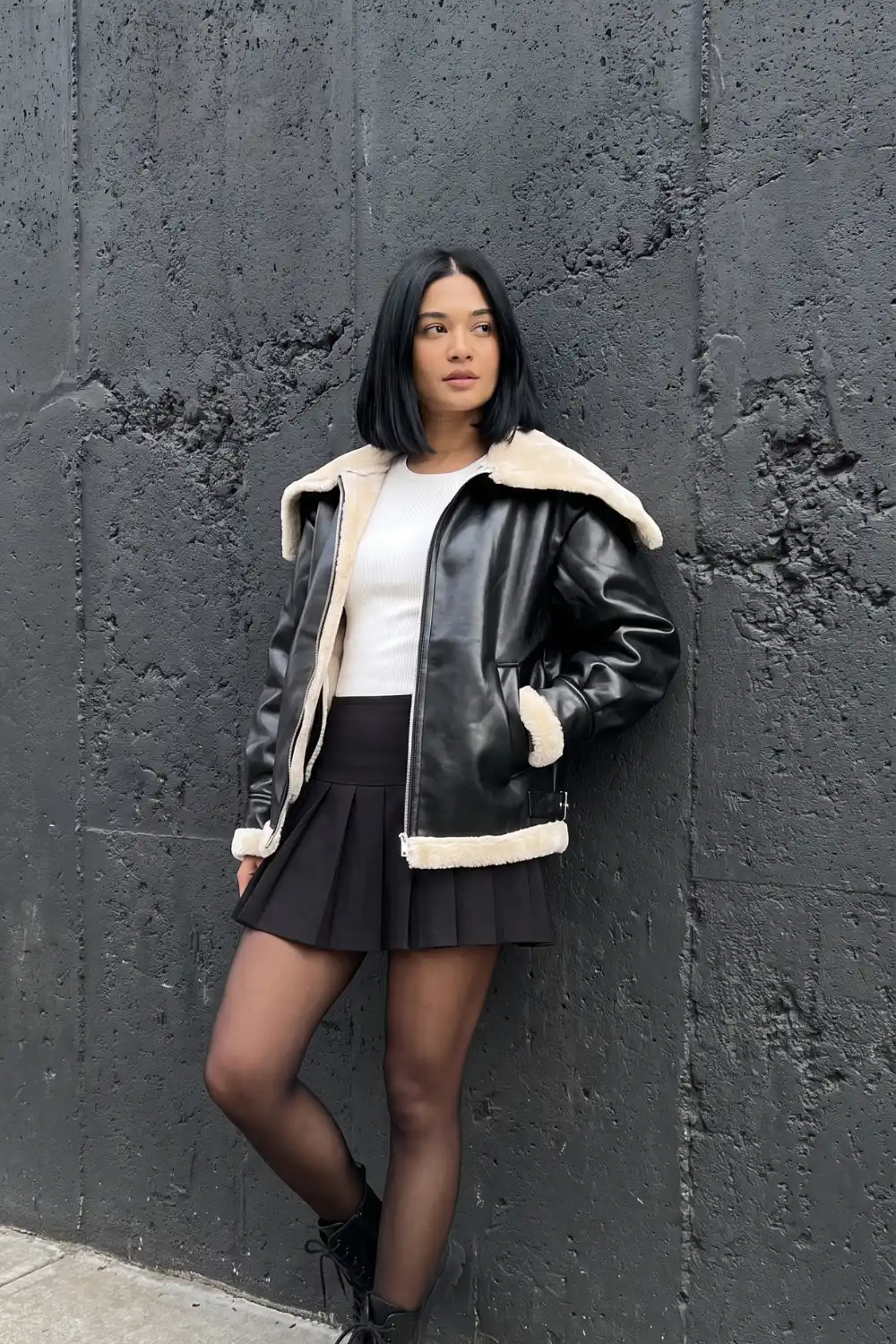 Woman wearing black faux leather shearling aviator jacket with white top black pleated mini skirt and combat boots