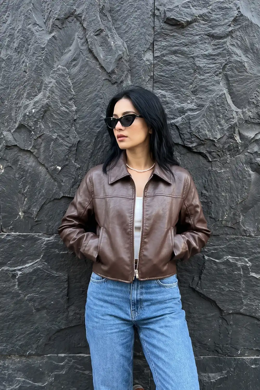 Woman wearing chocolate brown cropped zip up collared leather jacket with white crop top and light wash straight jeans