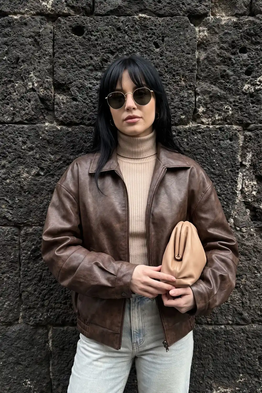 Woman wearing chocolate brown distressed leather bomber jacket with cream ribbed turtleneck and light wash jeans