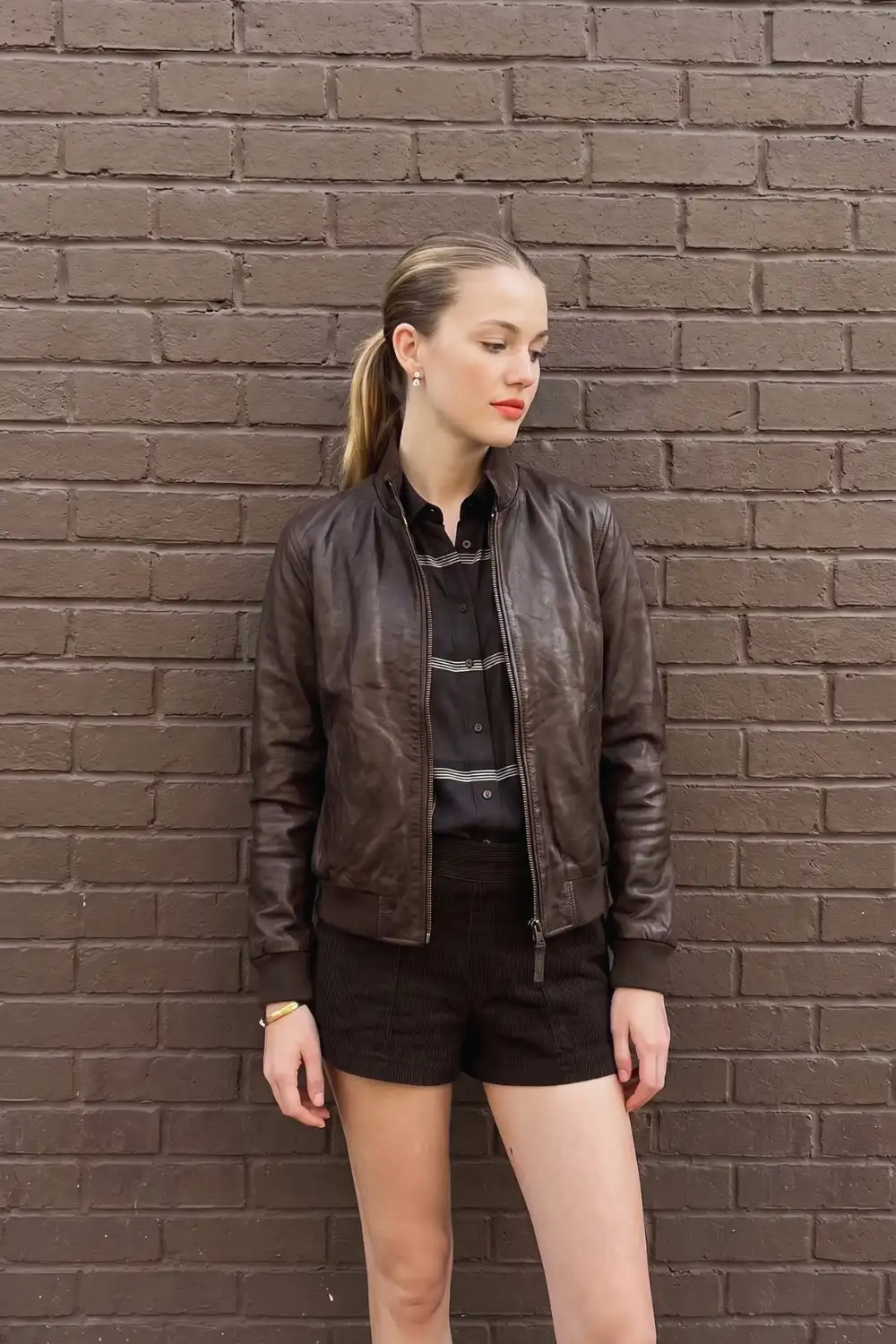 Woman in dark brown leather bomber jacket with black striped shirt and dark corduroy mini shorts