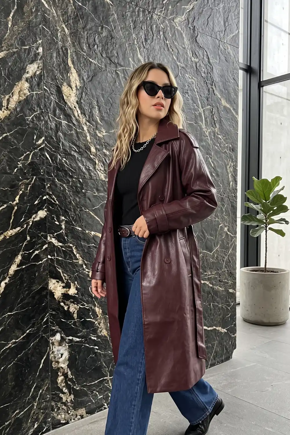 Woman wearing burgundy double breasted longline leather trench coat with black tee and dark wash wide leg jeans