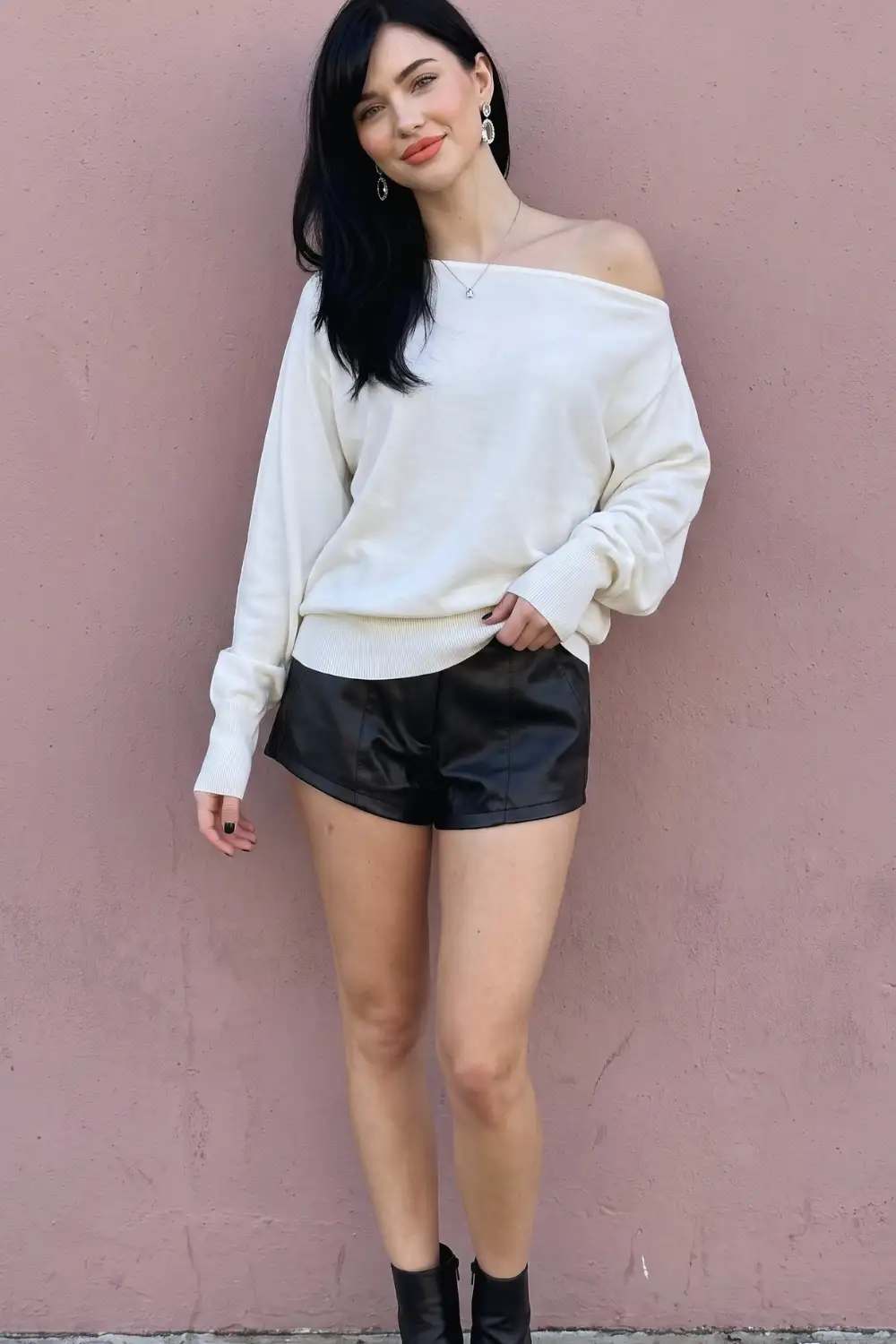 Woman in cream off shoulder oversized knit sweater with black leather mini shorts and ankle boots