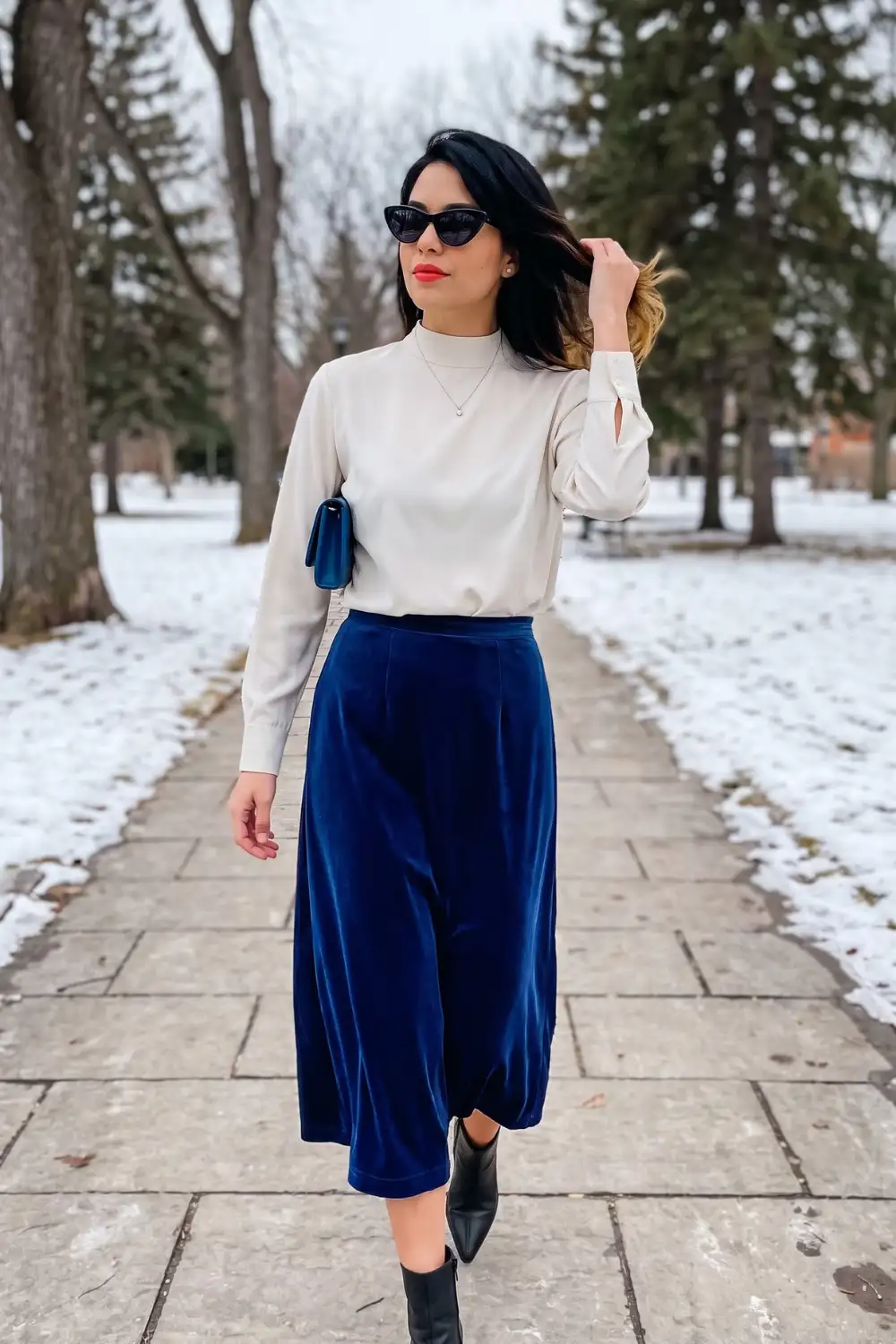 Woman in cream mock neck satin top with royal blue velvet midi skirt and black ankle boots