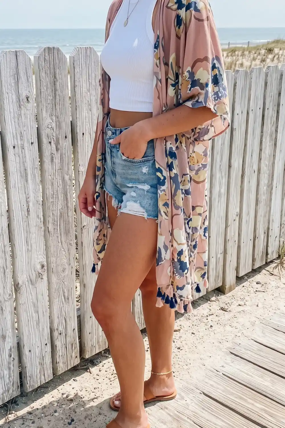 Woman wearing blush pink floral tassel kimono with white crop top and denim cutoff shorts at beach