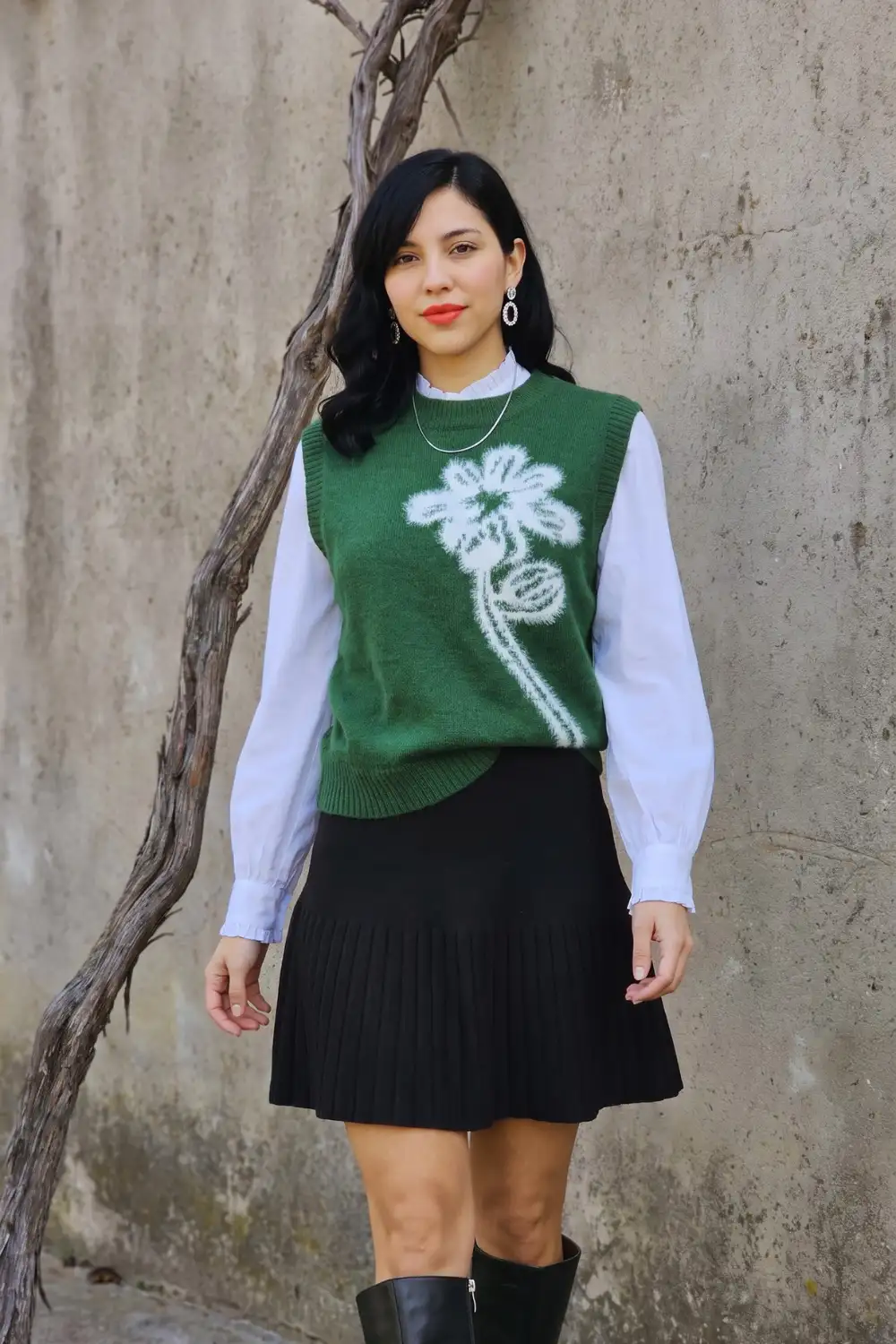 Woman in green floral intarsia knit vest over white ruffle shirt with black pleated mini skirt