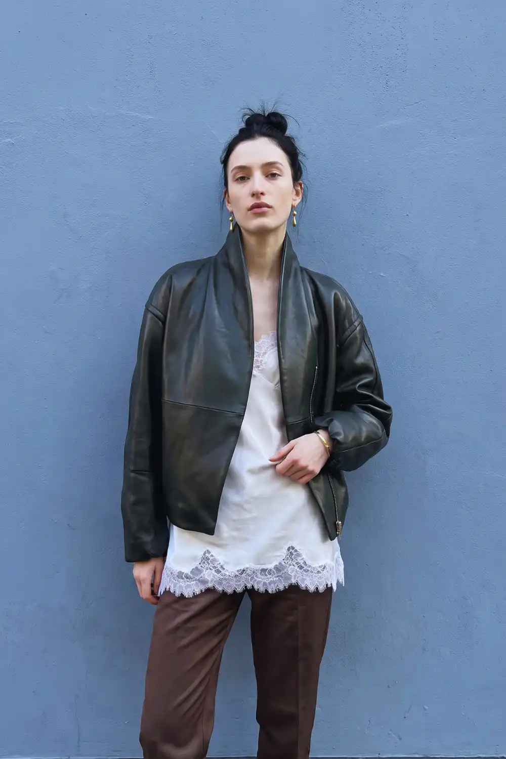 Woman in dark forest green oversized leather jacket with cream lace slip top and brown leather trousers