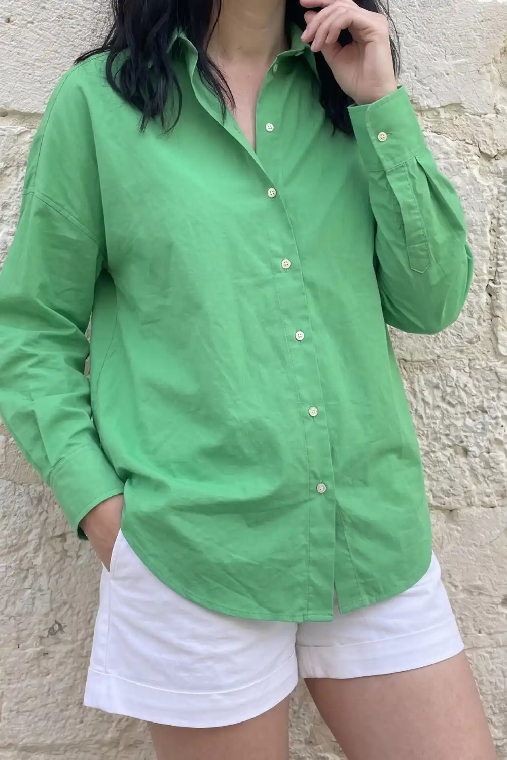 Woman wearing green oversized button-up shirt with white shorts against a stone wall