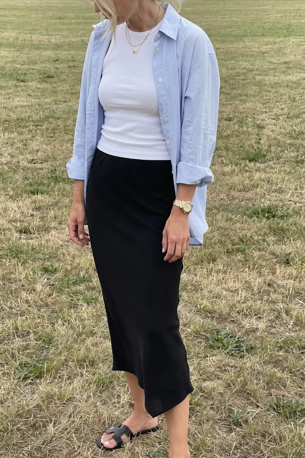 Woman wearing light blue oversized shirt over white ribbed tank top with black satin midi skirt