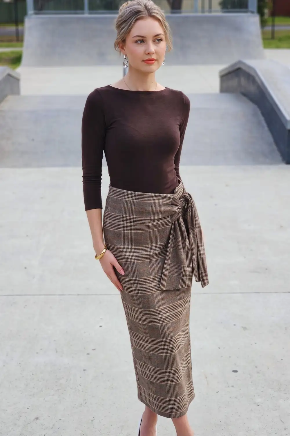 Woman in dark brown fitted bateau neck top with brown plaid wrap knot midi pencil skirt