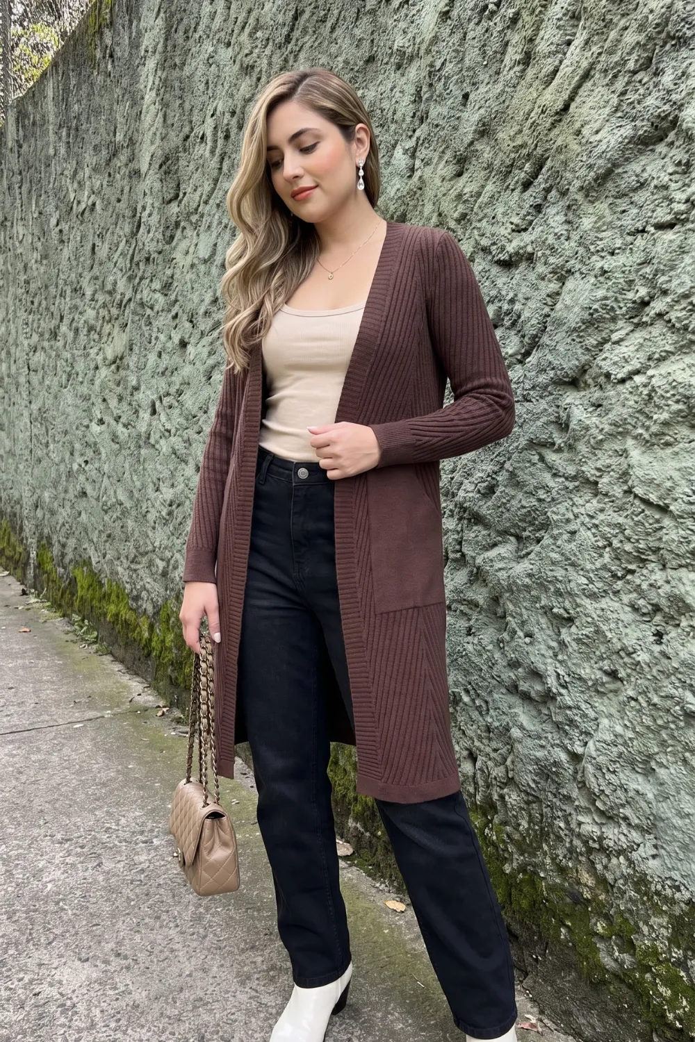 Woman in dark brown longline ribbed cardigan with beige tank and black straight jeans and white boots