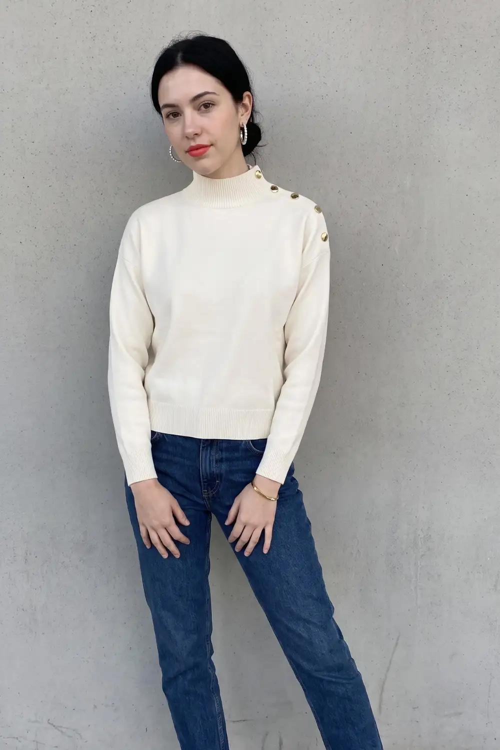 Woman in cream mock neck sweater with gold button shoulder detail and blue straight jeans