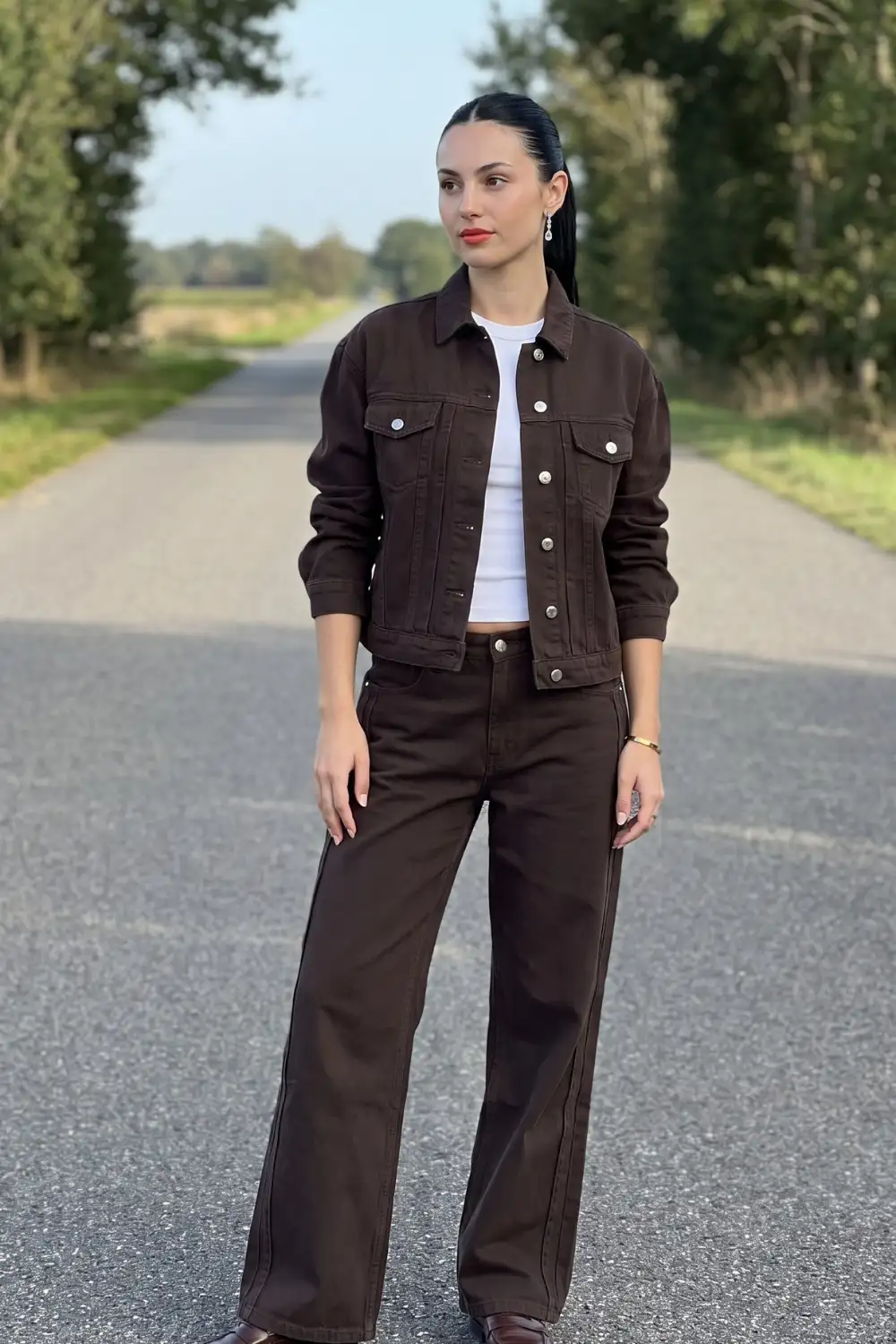 Woman in dark chocolate brown denim jacket with white crop top and matching wide leg jeans