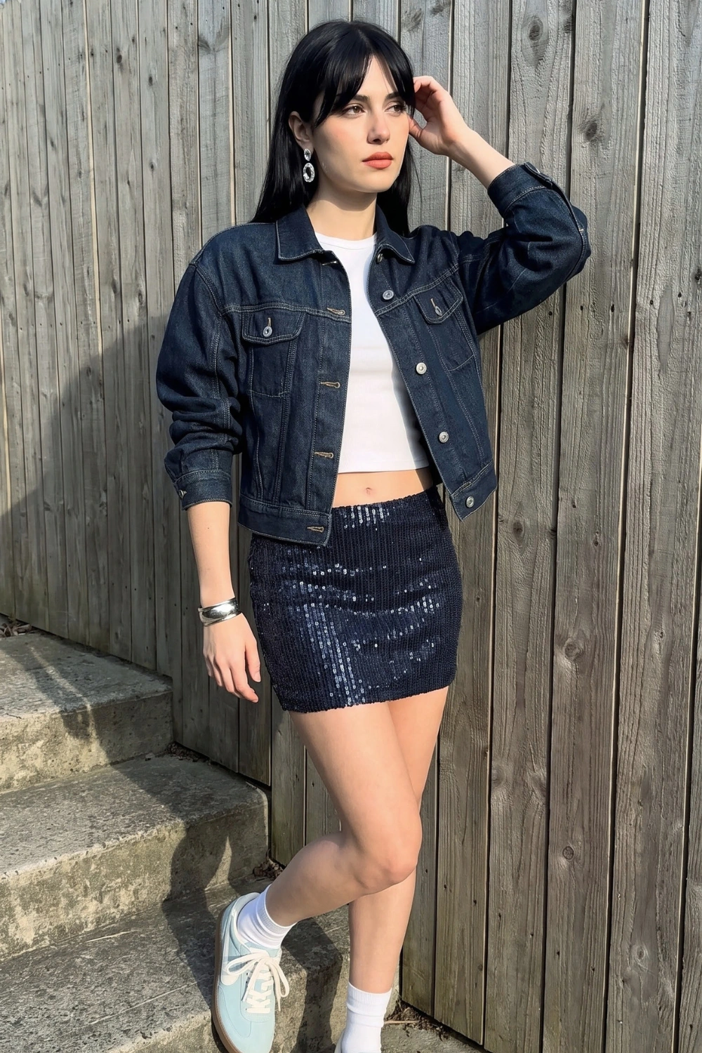 Woman in dark navy denim jacket with white crop top and navy sequin mini skirt and blue sneakers