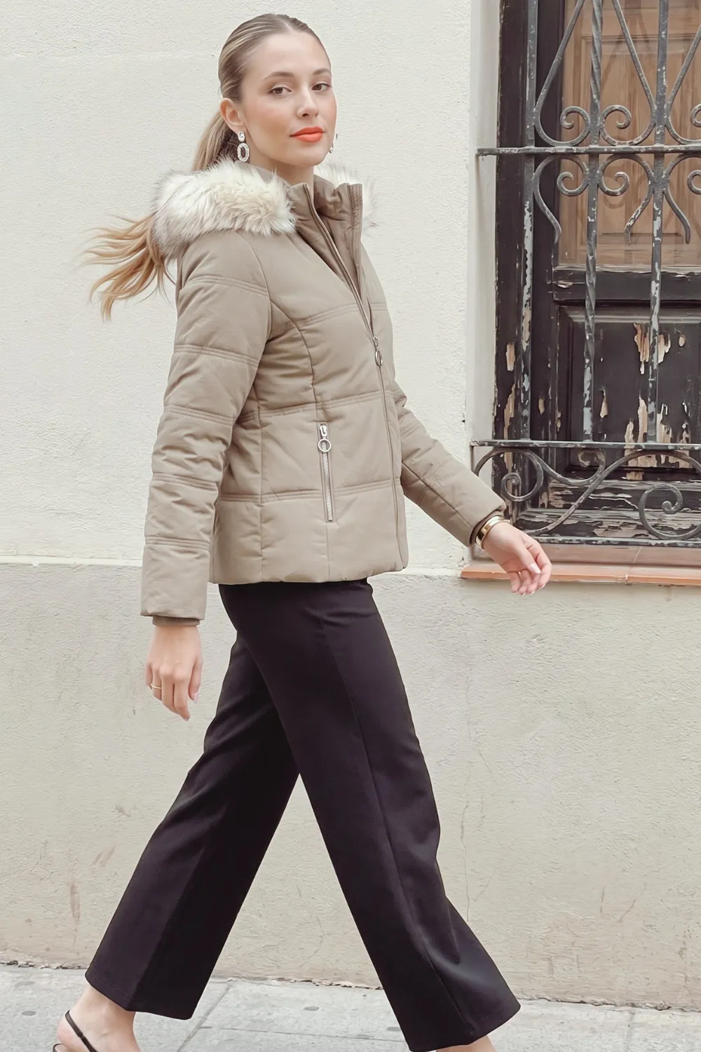Woman in taupe faux fur hood puffer jacket with black wide leg trousers
