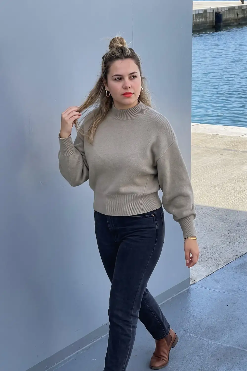 Woman in taupe oversized mock neck sweater with dark navy slim jeans and brown loafers